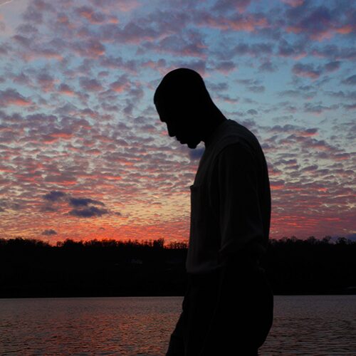 Silhouette standing near water, in front of clouds at sunset.