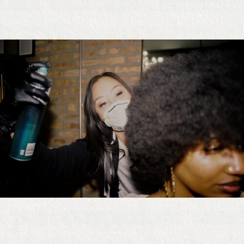 Image of an Asian stylist wearing a face mask, doing the hair of a Black model.