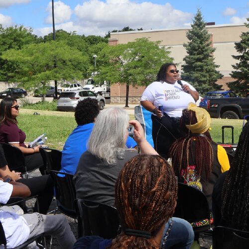 A community organizer speaks to a group, wearing a t-shirt that reads "Melcalf Park Proud".
